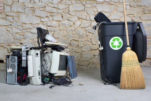 A pile of discarded electronic waste and broken computer hardware, including monitors, towers, cables, and internal components, stacked on a concrete surface in front of a textured stone wall. To the right, a large black rubbish bin with a green recycling symbol is positioned upright, accompanied by a traditional straw broom leaning against it. The scene suggests a clean-up or disposal area for electronic and general rubbish in a residential or commercial setting, with the environment characterized by natural light and an orderly arrangement of waste items ready for collection by a rubbish removal service such as Garden Clearance Docklands.