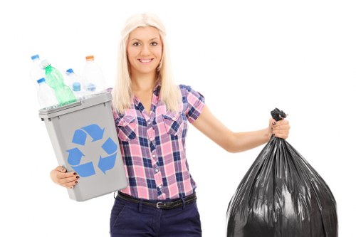 A young woman with long blonde hair, dressed in a short-sleeved, blue and pink checked shirt and dark trousers, stands against a plain white background. She is smiling and holding a small grey recycling bin filled with various plastic bottles, including clear, green, and blue types, positioned in her left hand. The recycling bin features a blue recycling symbol on the front. In her right hand, she holds a large black plastic rubbish bag, which appears to be filled and loosely tied at the top. The overall scene presents a clear, professional image emphasizing waste disposal and recycling, suitable for a rubbish removal service. This visual could relate to environmentally conscious rubbish collection, aligning with services offered by companies such as Garden Clearance Docklands in East London, supporting local waste management efforts.