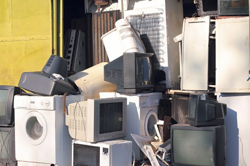 A collection of discarded household appliances and electronic waste situated outdoors, including a white washing machine, several CRT computer monitors with dark and light grey casings, and a black microwave oven stacked among other metal cabinets and broken electronic components. The appliances and debris are piled together on a paved surface, with some items leaning against each other and a white bucket placed atop a tall metal wire rack. The background consists of a partially open industrial storage area with yellow and grey metal doors, suggesting a waste disposal or recycling site. The scene depicts typical rubbish removal for electronic waste clearance from a property or commercial premises in the Docklands area, managed by Garden Clearance Docklands as part of their rubbish disposal services. Natural daylight highlights the textures of the various materials—smooth plastic, cold metal, and glass—emphasizing the mixed nature of disposed electronic and household waste awaiting removal and proper disposal.