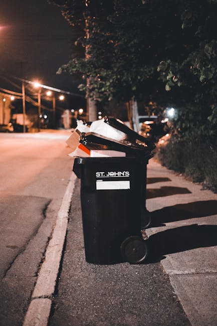 A black wheeled rubbish bin positioned on the edge of a paved sidewalk in a residential area at night, with the label 'ST. JOHN'S' visible on its side. The bin is overflowing with mixed waste, includi