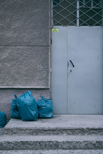 A black wheeled rubbish bin positioned on the edge of a paved sidewalk in a residential area at night, with the label 'ST. JOHN'S' visible on its side. The bin is overflowing with mixed waste, includi
