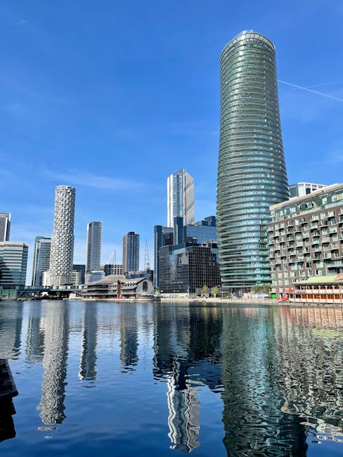 A cityscape featuring a variety of modern high-rise buildings along a waterfront, with the tallest structure being a cylindrical glass tower with horizontal lines running around its exterior. Several 